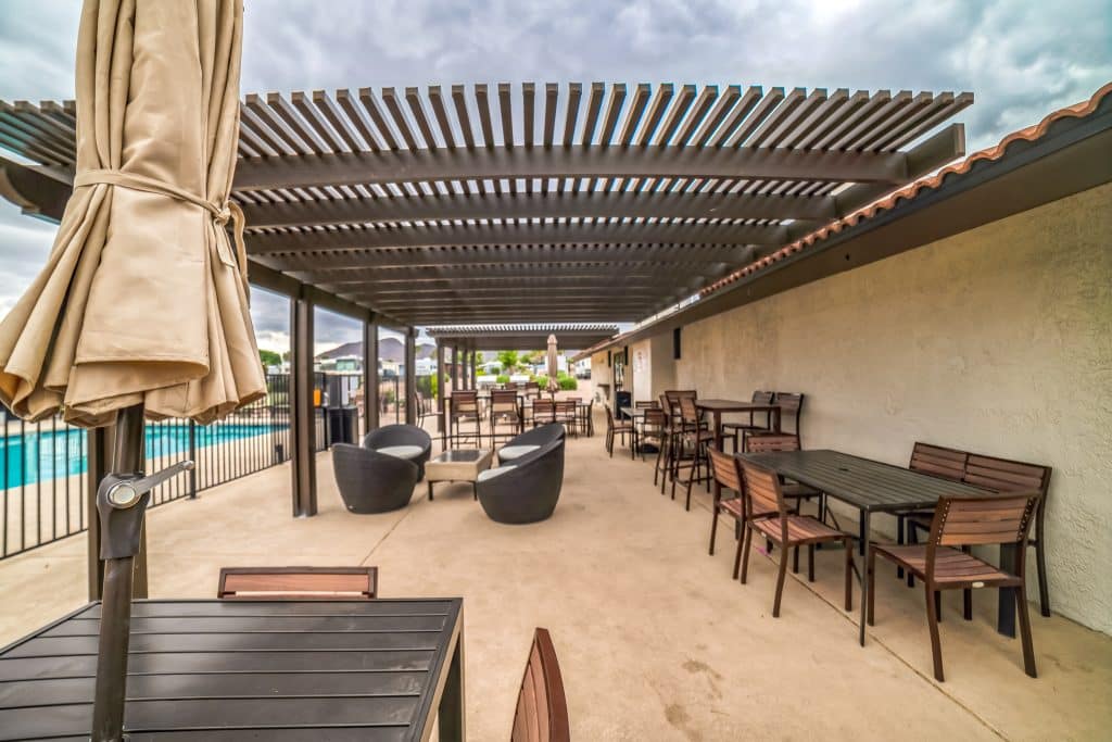 Outdoor covered patio with a pergola, featuring black tables, wooden chairs, and cushioned seats. The patio overlooks a pool and is surrounded by fencing in henderson nv